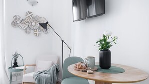 A modern living room with a round wooden table, a white armchair, a floor lamp, a TV mounted on the wall, and a decorative wall piece.