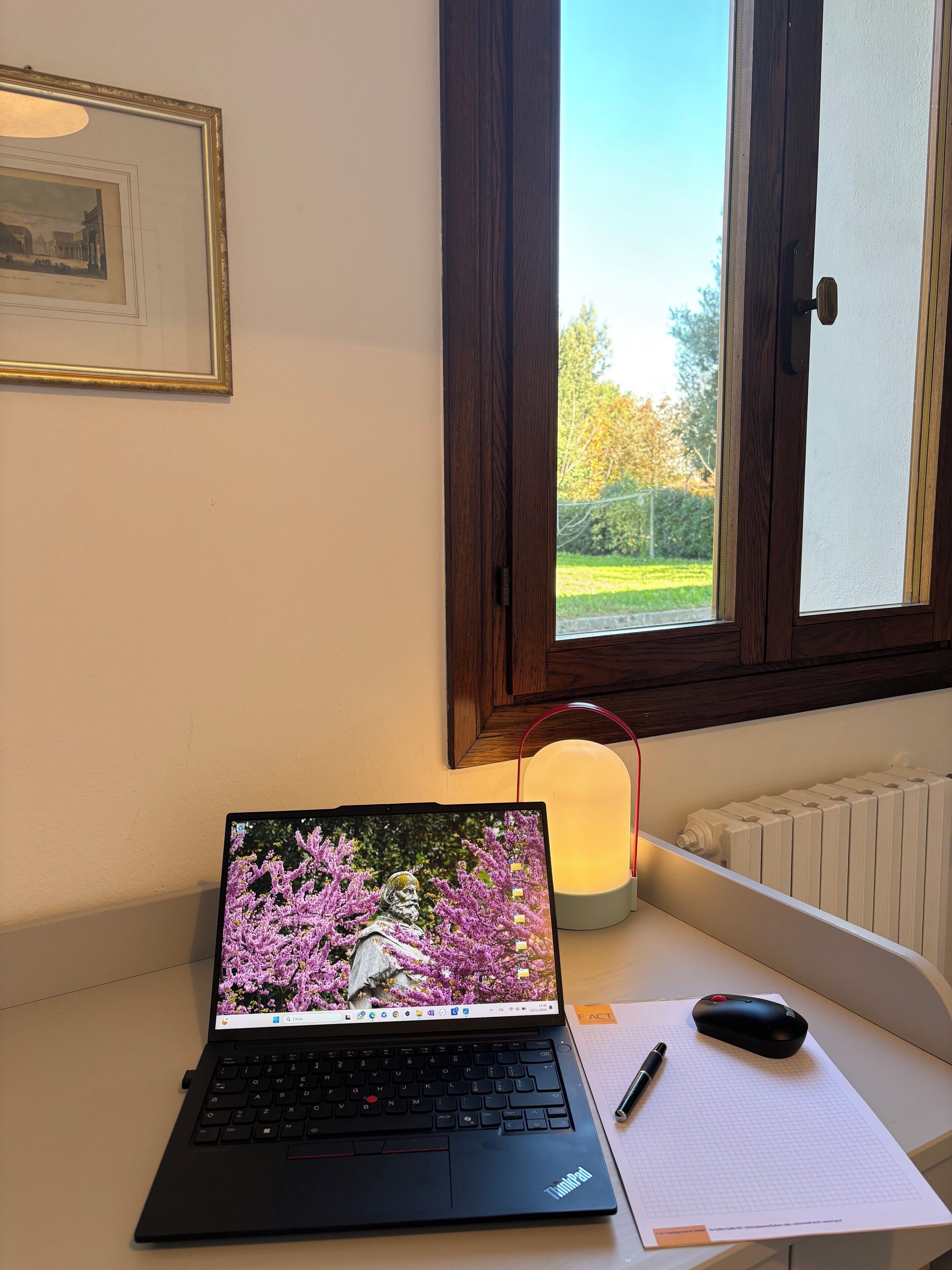 A laptop on a desk with a mouse, a pen, and a notepad. There is a window with a view of greenery outside.