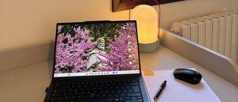 A laptop on a desk with a mouse, a pen, and a notepad. There is a window with a view of greenery outside.