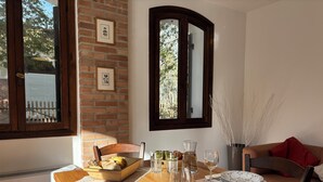 Un coin repas avec une table en bois dressée pour deux personnes, un mur de briques orné de tableaux, et une fenêtre donnant sur des arbres et une clôture.