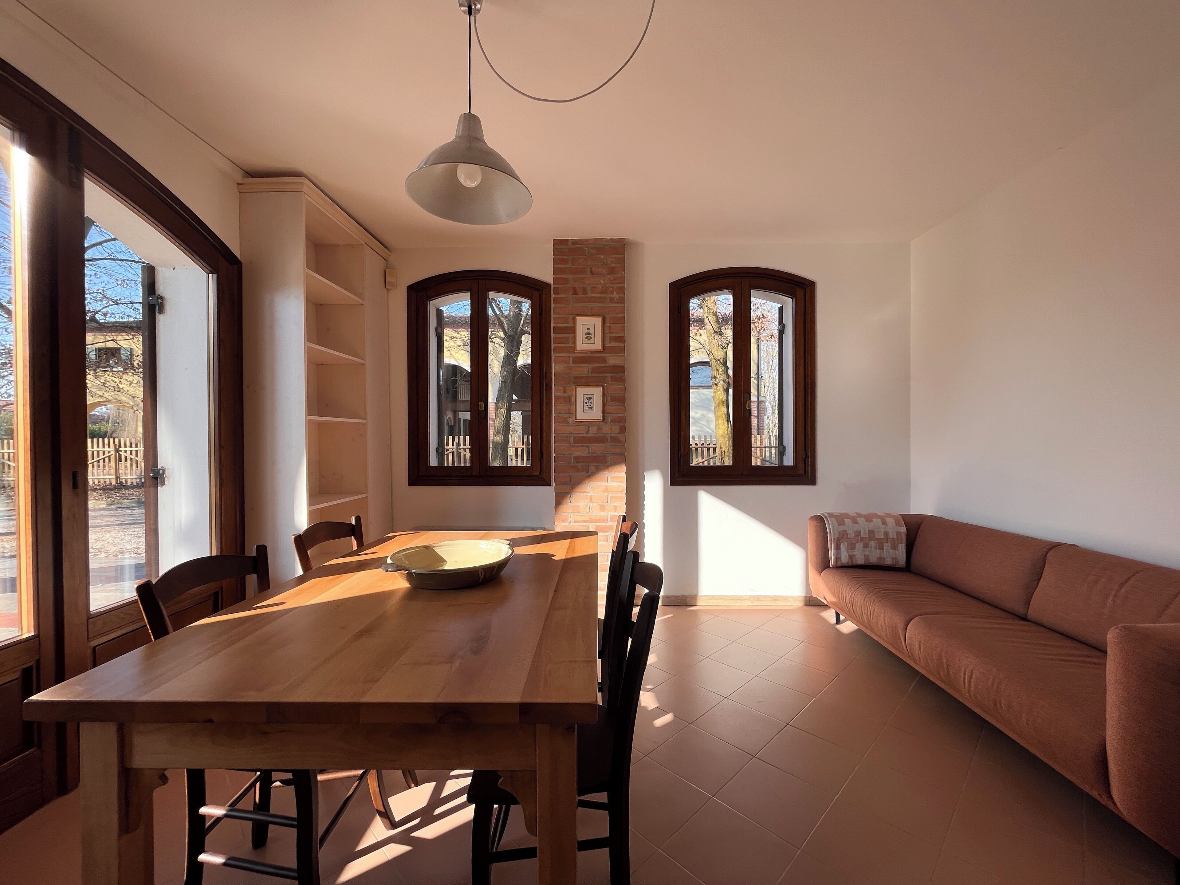 Un coin repas avec une table et des chaises en bois, une grande fenêtre donnant sur un mur de briques et un canapé marron dans l’espace salon.