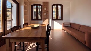 Un coin repas avec une table et des chaises en bois, une grande fenêtre donnant sur un mur de briques et un canapé marron dans l’espace salon.