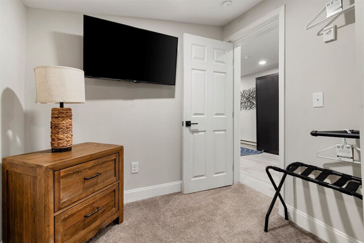 A room with a wooden dresser, a flat-screen TV mounted on the wall, a white door, and a black metal rack.