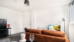 Una sala moderna con un sofá marrón, una mesa de comedor con platos y una copa de vino, un televisor en la pared y un cubo de basura.