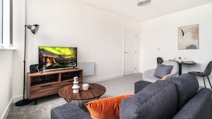 A modern living room with a flat-screen TV, wooden entertainment unit, grey sofa, orange cushion, and a dining table with chairs. - Fabulous 1 BED Apartment Central Wrexham with WiFi (Wrexham)