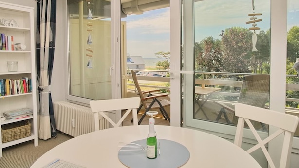 Une pièce avec une grande fenêtre, une table sur laquelle se trouve une bouteille, une chaise, une bibliothèque et un radiateur.