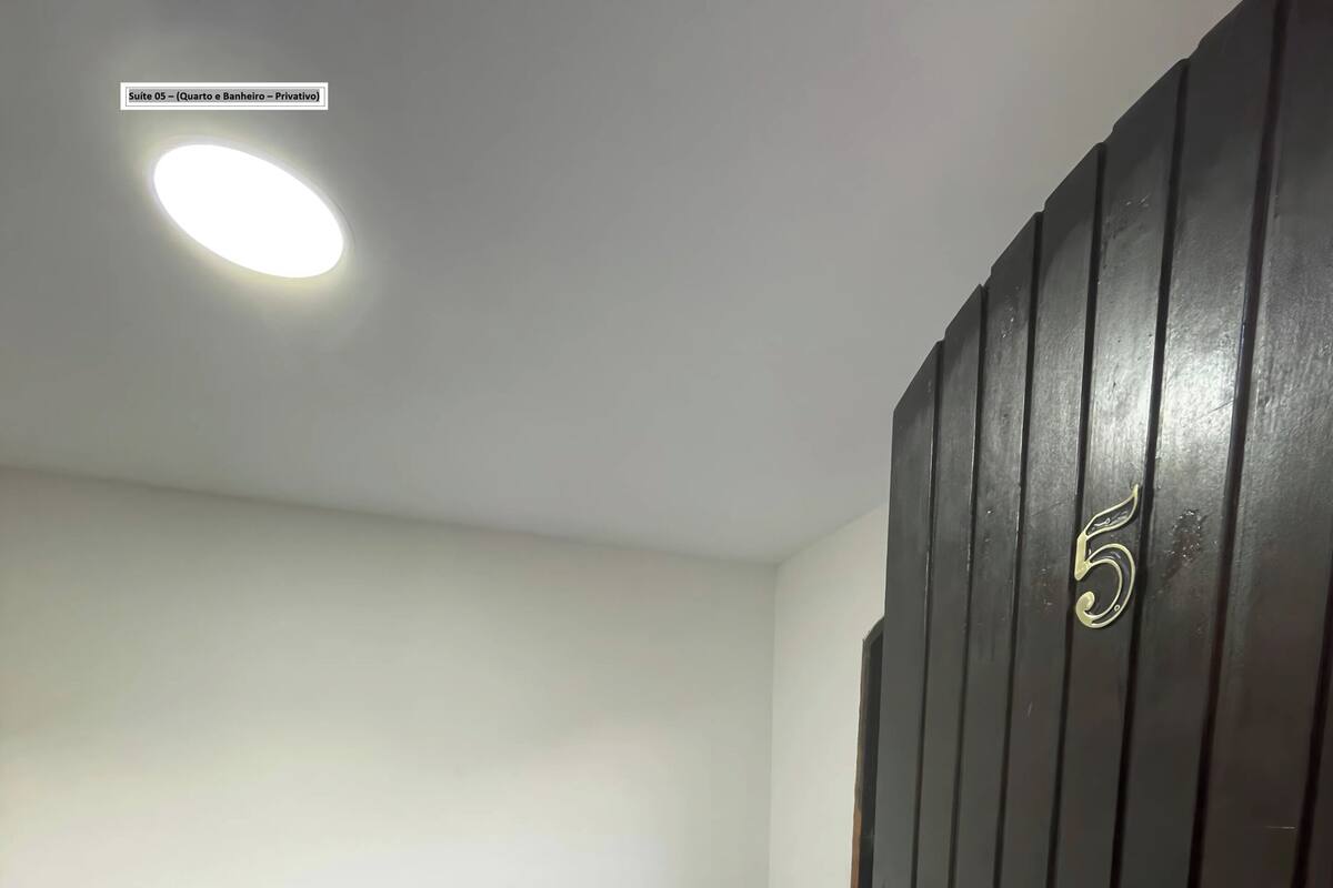 Een kleine kamer met een enkel bed, een blauwe matras, een witte muur, een zwarte deur met het nummer 5 en een plafondlamp.