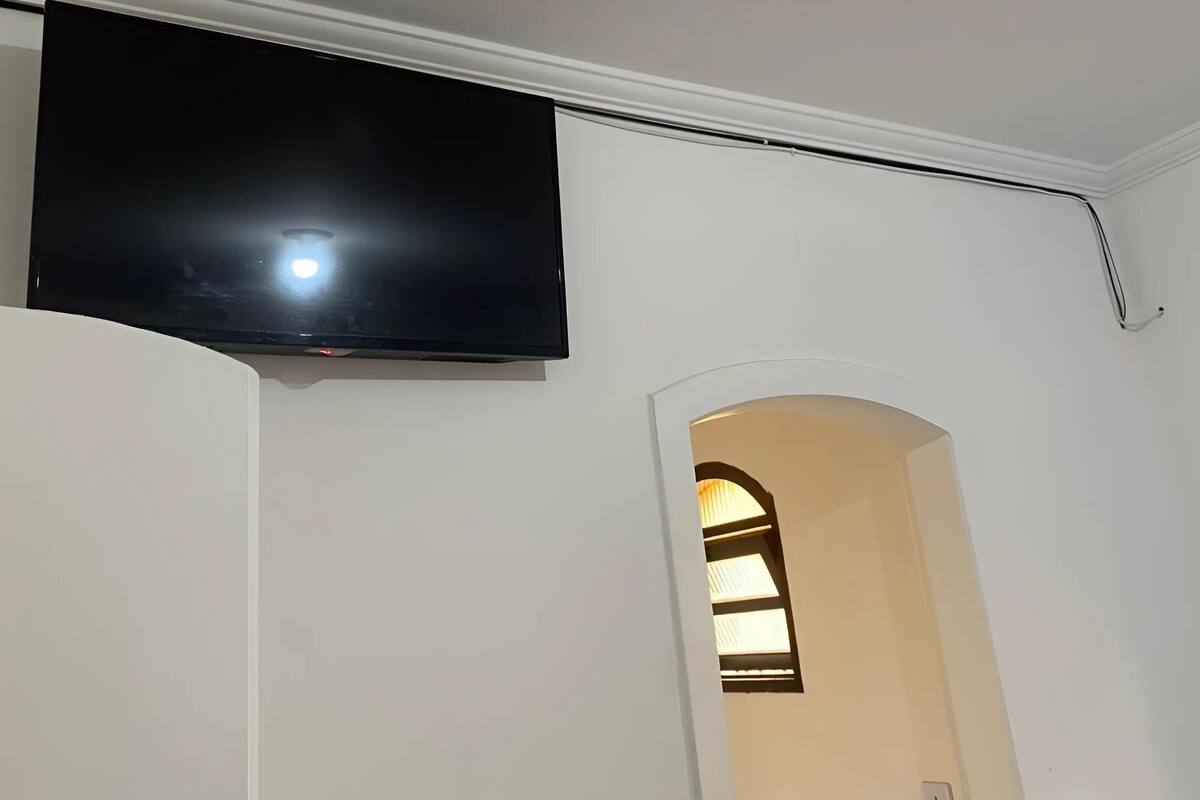 Een kamer met een wandgemonteerde flatscreen televisie, een doorgang naar een andere kamer, en een wit plafond.