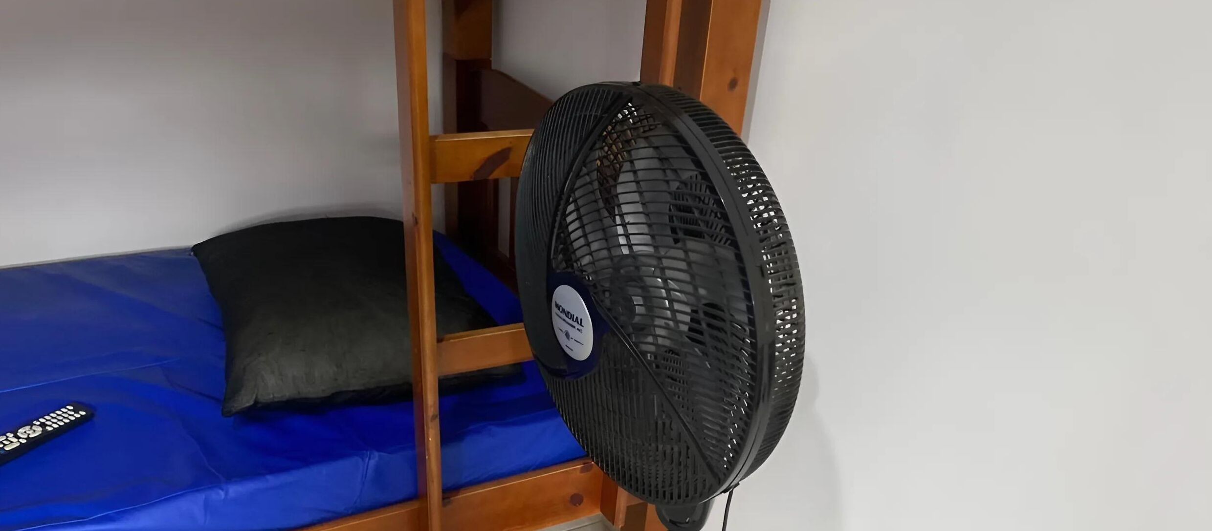Uma cama beliche com colchão azul e travesseiro preto. Um ventilador preto com grade circular está posicionado na parede.