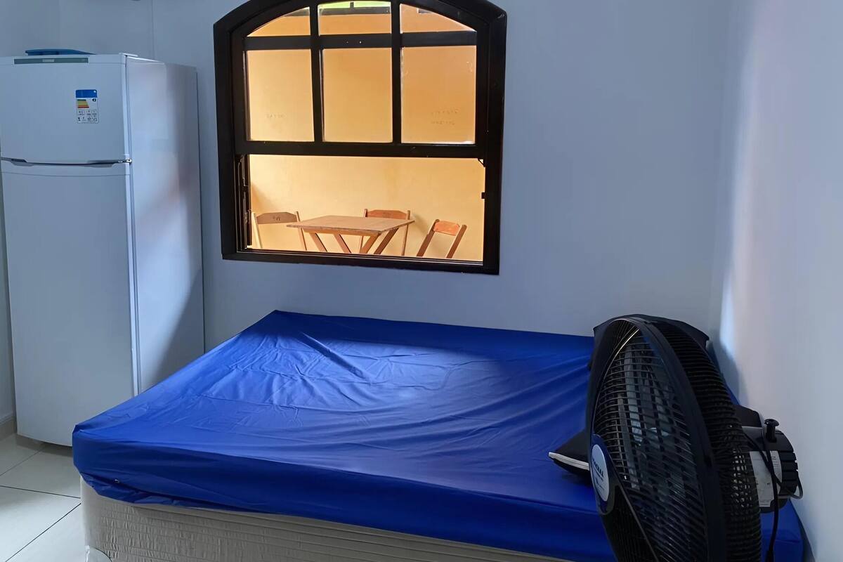 Een kleine kamer met een bed, een raam, een ventilator en een koelkast.
