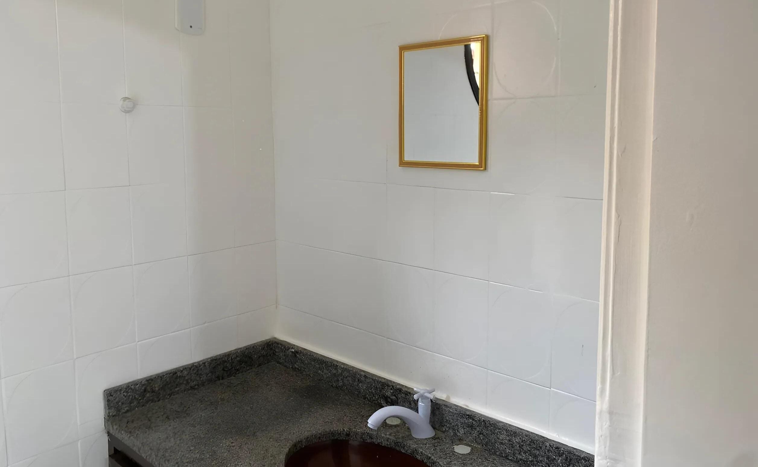 Casa de banho com paredes de azulejos brancos, bancada de granito, torneira branca e espelho com moldura dourada.