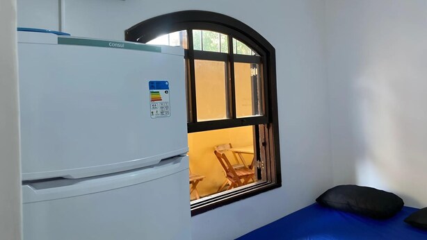 Une pièce de taille modeste, avec un réfrigérateur blanc, une fenêtre à cadre noir, un lit bleu et une chaise en bois, visibles à travers la fenêtre.