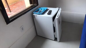 Une petite machine à laver blanche, avec un bac bleu sur le dessus, une télécommande et une fenêtre donnant sur un sol recouvert de carreaux rouges.