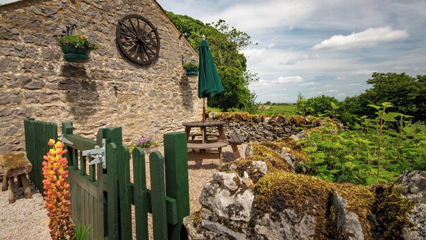 A rustic stone building with a wooden wheel, a green umbrella, and a wooden bench. - Bolehill Farm Cottages (Bakewell)