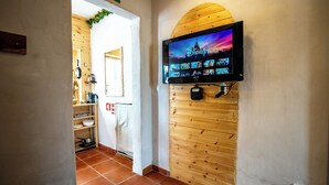 Un pasillo con pared de madera, un televisor montado que muestra un canal de Disney y una zona de cocina con estantes y una cafetera.