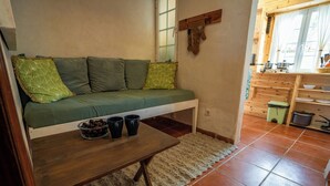 Una zona de estar acogedora con un sofá, una mesita con un cuenco de fruta y tazas, y una cocina con armarios de madera y un ventanal.