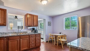 A kitchen with wooden cabinets, granite countertops, a stainless steel refrigerator, and a dining area with wooden chairs. - Sonoran Velo Summary: (Tucson)