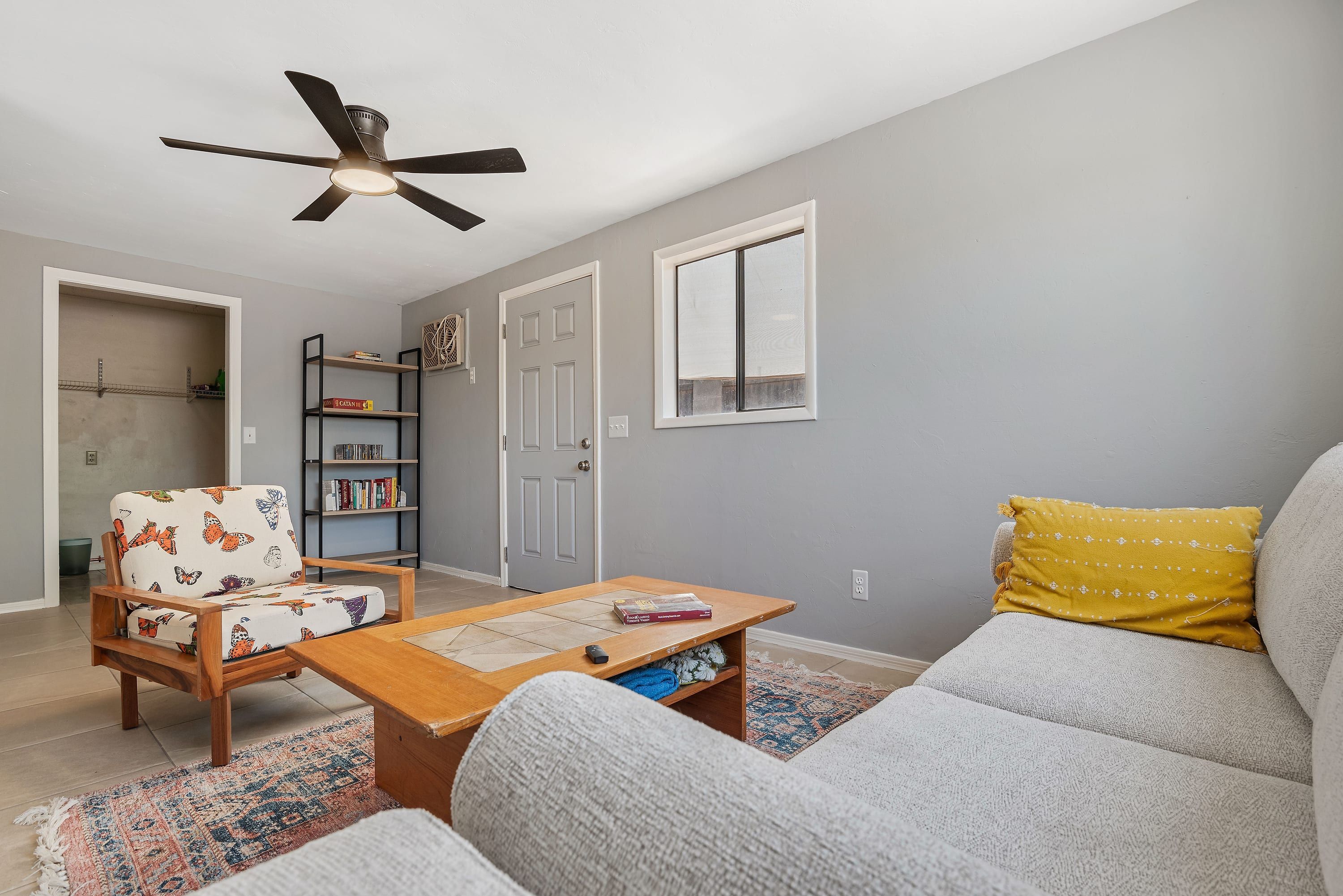 A living room with a ceiling fan, a sofa, a chair with a butterfly pattern, a coffee table, a bookshelf, and a door.