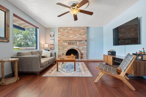 A living room with a brick fireplace, a ceiling fan, a sofa, a coffee table, and a TV mounted on the wall. - Sonoran Velo (Tucson)