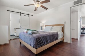 A bedroom with a wooden bed, a ceiling fan, a white door, and a nightstand with a lamp. - Sonoran Velo (Tucson)