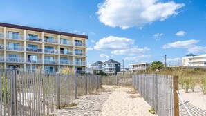 A sandy pathway leading to a multi-story building with balconies, surrounded by a wooden fence. - Direct Oceanfront with Pool - Steps To The Beach (Ocean City)
