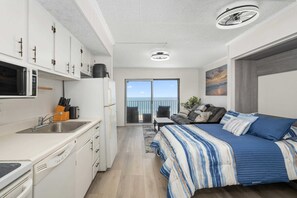 Ein kompakter, moderner Raum mit Küchenzeile, einem Bett mit gestreifter Bettwäsche, Blick durch eine Glastür nach draußen und einem Deckenventilator.