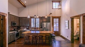 A spacious kitchen with wooden cabinets, a large island with stools, hanging pendant lights, and a view through the windows. - Doe Run Cottage at Eagles Nest (Banner Elk)