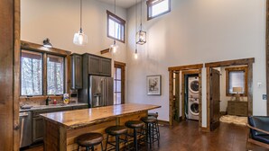 A spacious kitchen with a large island, bar stools, and wooden cabinetry. There are hanging lights and a view of the outdoors through windows. - Doe Run Cottage at Eagles Nest (Banner Elk)