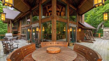 A wooden deck with a round table and benches, surrounded by glass doors and windows, overlooking a forested area.