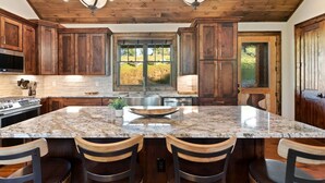 A kitchen with wooden cabinets, a large island with bar stools, a stainless steel sink, and a view through the window. - Timberwolf Lodge at Eagles Nest (Banner Elk)