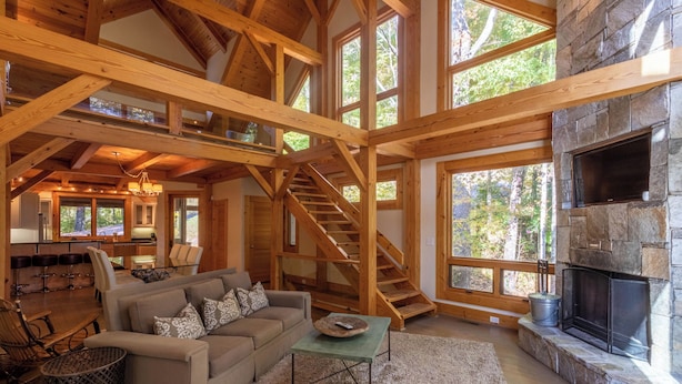 A spacious living area with a stone fireplace, wooden beams, a staircase, and large windows. - Modern Cedar Rock Townhome at Eagles (Banner Elk)