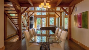 A dining area with a glass-top table and leather chairs, wooden beams, a chandelier, and large windows with a view of trees. - Modern Cedar Rock Townhome at Eagles (Banner Elk)