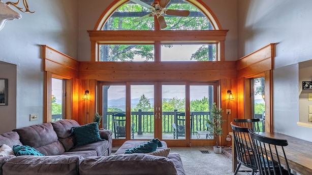 A spacious living room with a large wooden-framed window, a ceiling fan, and a view of a deck and mountains. - Mount Ever-Rest (Banner Elk)