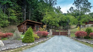 A wooden cabin with a gravel driveway leading to a gated entrance, surrounded by a forested area with various shrubs and trees. - Hilltop Hideout at Eagles Nest (Banner Elk)
