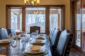 A dining room with a long wooden table set for six, surrounded by leather chairs, with a view of trees through large windows. - Fern Ridge Lodge at Sweetgrass (Blowing Rock)