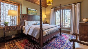 Ein Bett mit Baldachin, ein hölzernes Kopfteil, ein großes Fenster mit Jalousien, ein gemustertes Teppich und ein Nachttisch mit Lampe.