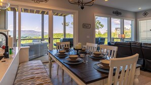 Uma sala de jantar com uma mesa grande posta para quatro pessoas, rodeada por cadeiras brancas, com vista para o exterior.