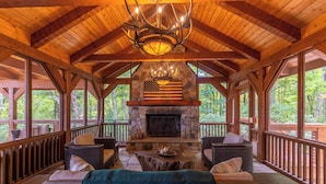 A covered outdoor seating area with stone fireplace, wooden beams, and wicker furniture. - Matney Ridge Estate (Banner Elk)