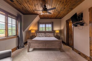 Quarto com teto de madeira, cama com cabeceira, duas luminárias de mesa, televisão fixada na parede e janela com cortinas.