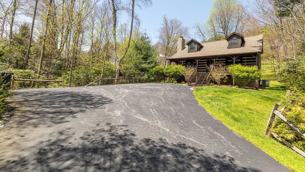 A log cabin with a stone chimney, surrounded by a forested area with a paved driveway leading to the entrance. - Hemlock Hideaway (Boone)