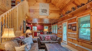A cozy cabin interior with a wooden staircase, a living room with two sofas and a coffee table, a kitchen area, and large windows with blinds. - Crosswind (Vilas)