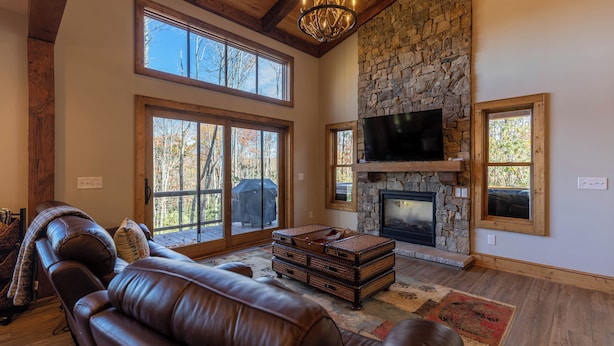 A spacious living room with a stone fireplace, leather sofas, and large windows offering a view of the outdoors. - Hidden Gem at Eagles Nest (Banner Elk)