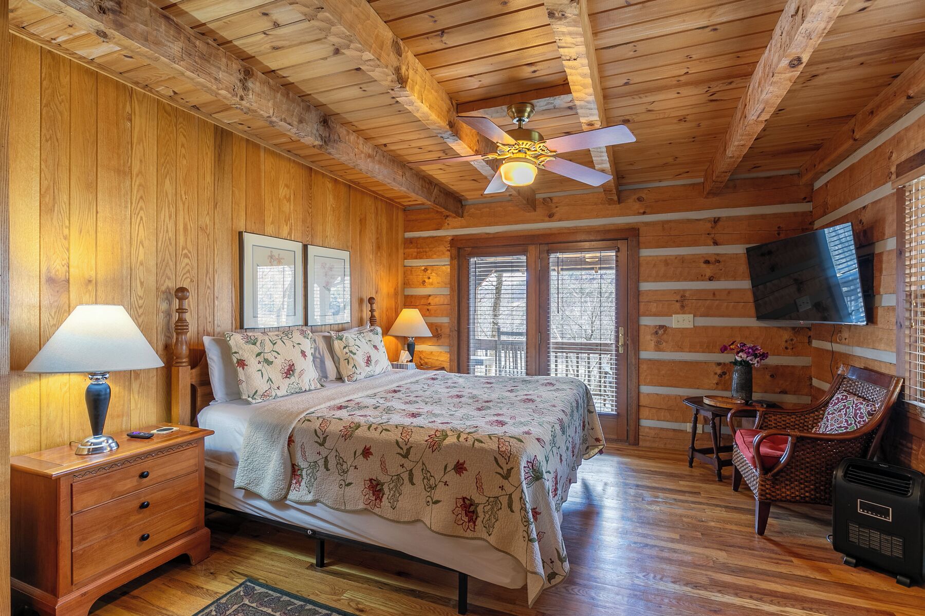 A cozy log cabin bedroom with a wooden ceiling, a bed with floral bedding, a nightstand, a chair, a television, and a window with blinds.