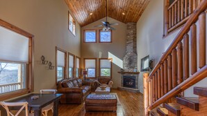 A spacious living room with a wooden ceiling, a stone fireplace, and a staircase with wooden railings. - Skyland Cabin (Boone)