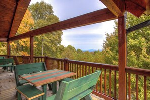 A wooden deck with green benches and a table, overlooking a forested area with trees and mountains in the distance. - The Nest at Seven Devils (Banner Elk)