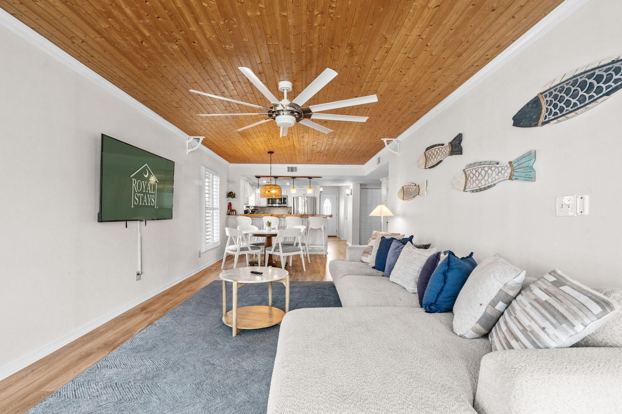 A modern living room with a ceiling fan, a dining area with white chairs, and a kitchen in the background.