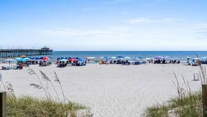 Een strand met parasols en ligbedden, een steiger op de achtergrond, en heldere luchten.