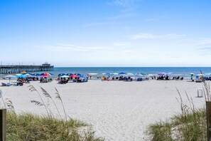 A beach with umbrellas and beach chairs, a pier in the background, and clear skies.