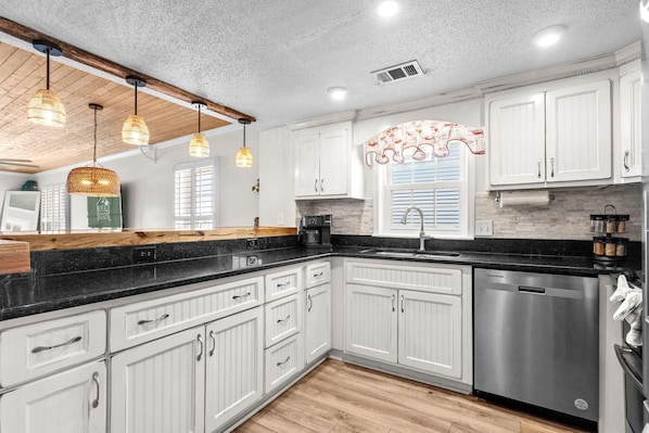A kitchen with white cabinets, a black countertop, a stainless steel dishwasher, and pendant lights.