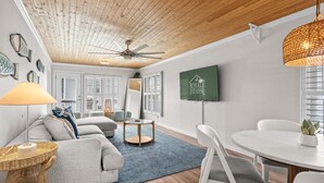 Een moderne woonkamer met een plafondventilator, een aan de muur bevestigde televisie en een eethoek met een tafel en stoelen.